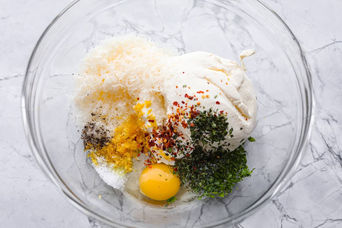 Irresistible Baked Ricotta Dip is the perfect party appetizer! It takes just minutes to combine creamy ricotta cheese with parmesan, fresh herbs, and lemon zest. Then, bake until bubbly and delicious, and get ready for an explosion of flavor! Overhead shot of dip ingredients in a glass bowl.