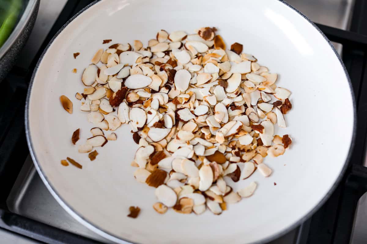 Green Beans Almondine is made with fresh green beans that are sautéed until crisp-tender and then tossed with buttery, toasted almonds. Every bite is filled with nutty richness and a hint of lemon to brighten up the flavors. Overhead shot of almonds toasting in a skillet.