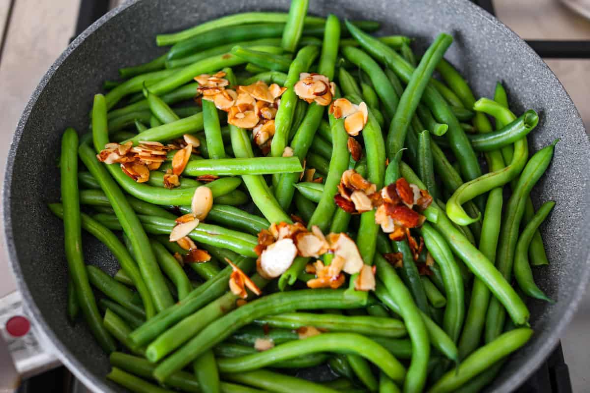 Green Beans Almondine is made with fresh green beans that are sautéed until crisp-tender and then tossed with buttery, toasted almonds. Every bite is filled with nutty richness and a hint of lemon to brighten up the flavors. Overhead shot of steamed green beans with the almond, butter and lemon juice poured over the top.