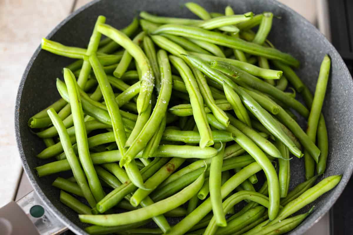 Green Beans Almondine is made with fresh green beans that are sautéed until crisp-tender and then tossed with buttery, toasted almonds. Every bite is filled with nutty richness and a hint of lemon to brighten up the flavors. Overhead shot of green beans, salt and water in a skillet.