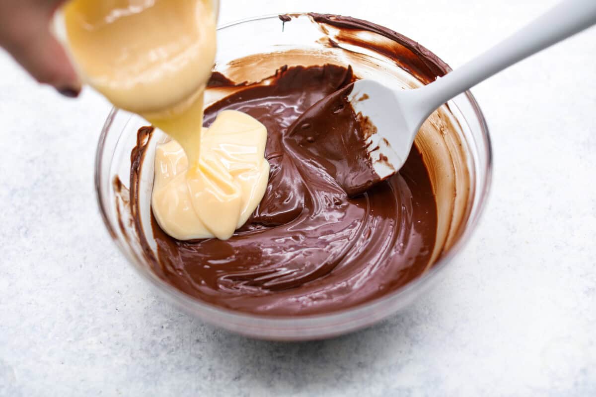 Overhead shot of someone pouring sweetened condensed milk into. the melted chocolate and butter mixture. 