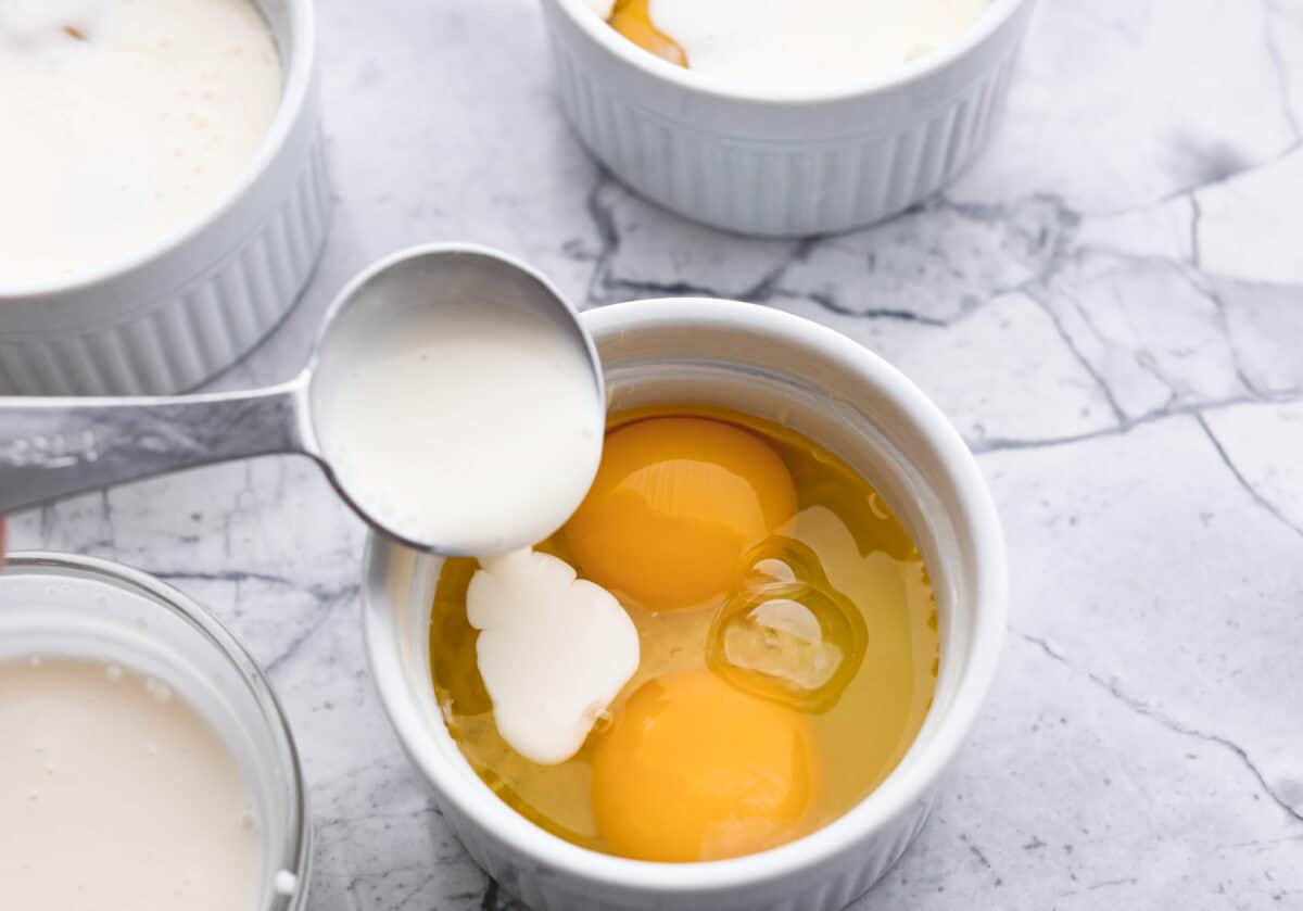 Photo of a ramekin with two eggs and someone pouring cream into it.