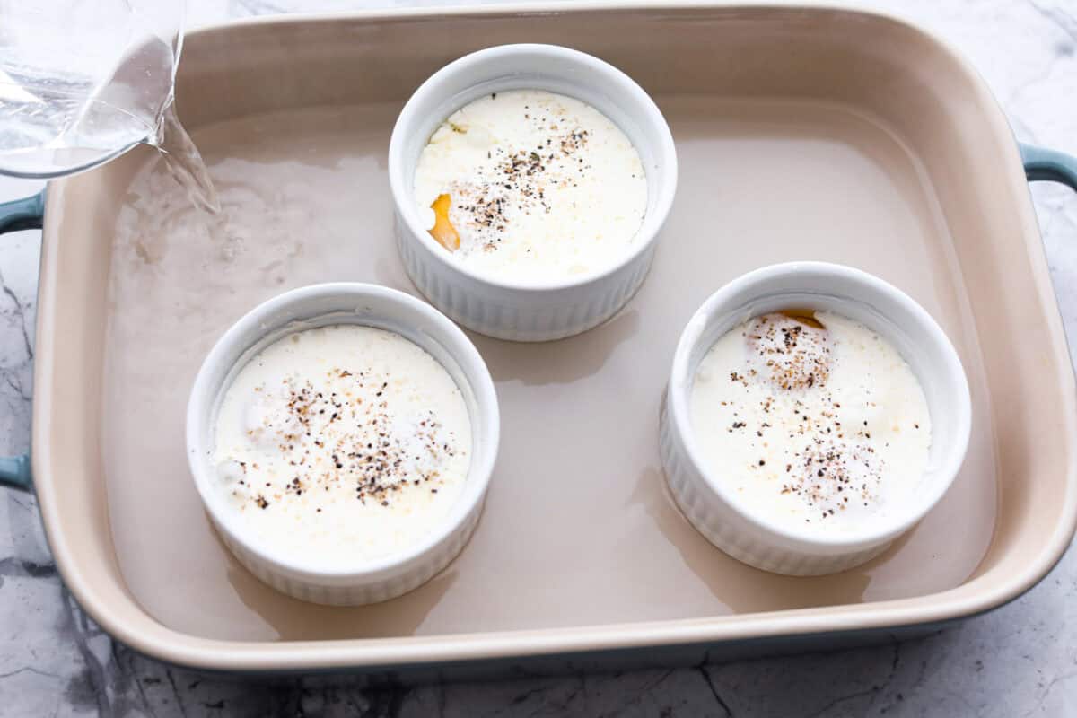 Overhead shot of three ramekins in baking dish with water being poured into the baking dish surrounding the ramekins. 