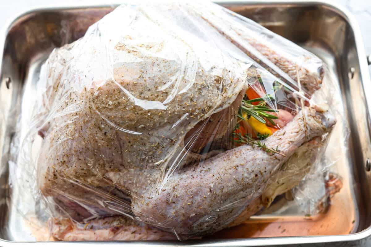 Side shot of the prepared turkey covered in plastic wrap in a pan. 