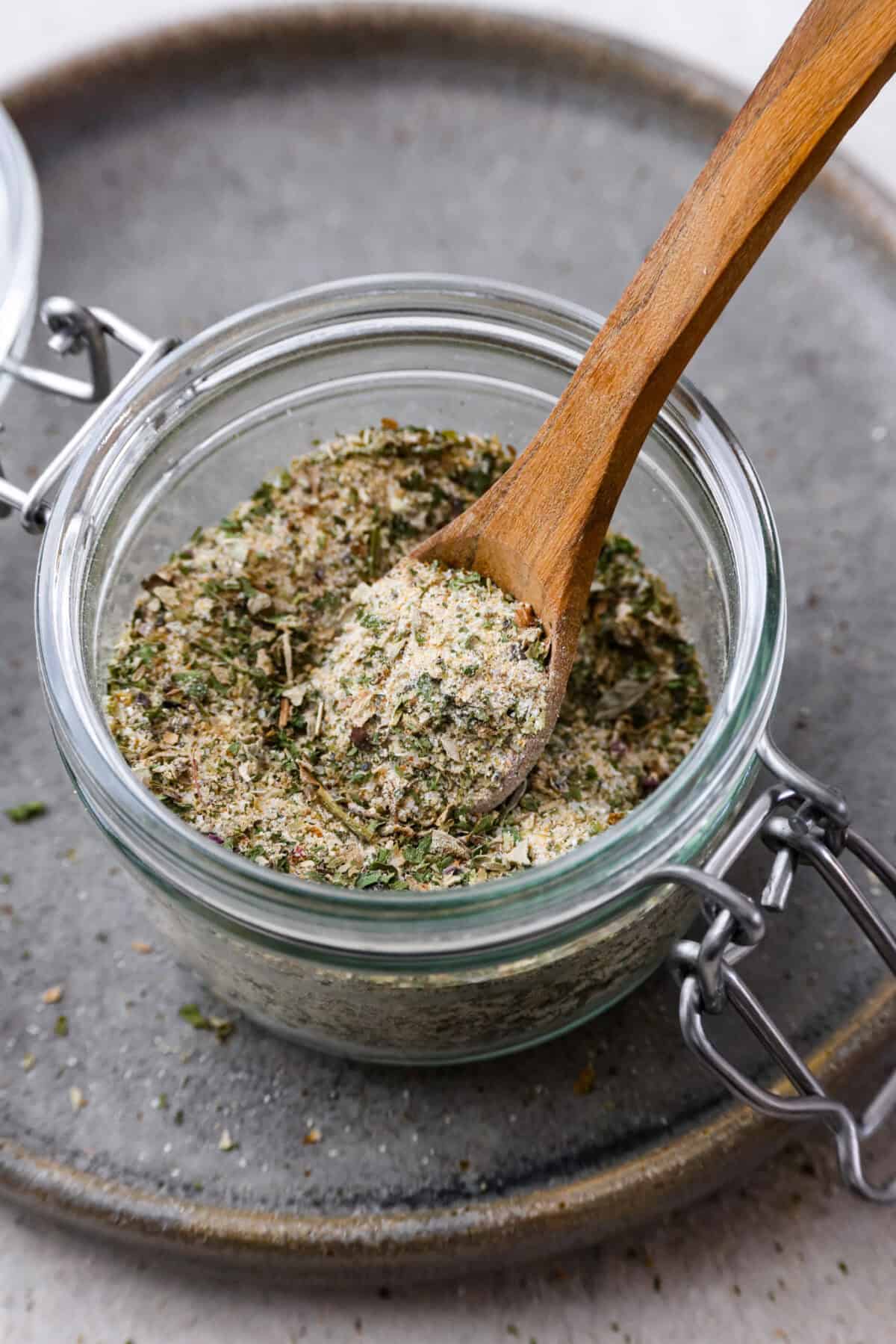 Angle shot of homemade garlic herb seasoning in a glass jar with a small wooden spoon sticking out. 