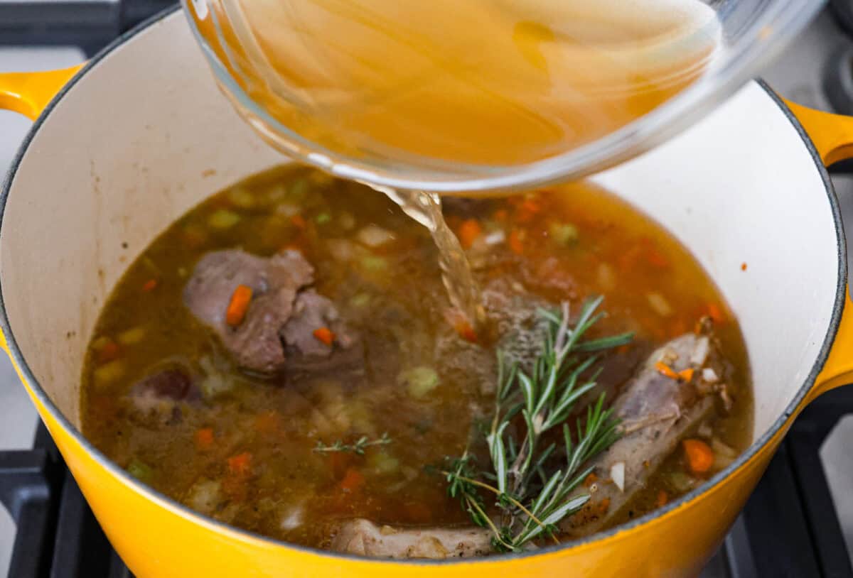 Angle shot of someone pouring chicken broth into the pot with giblets, vegetables, garlic and sprigs of fresh herbs. 