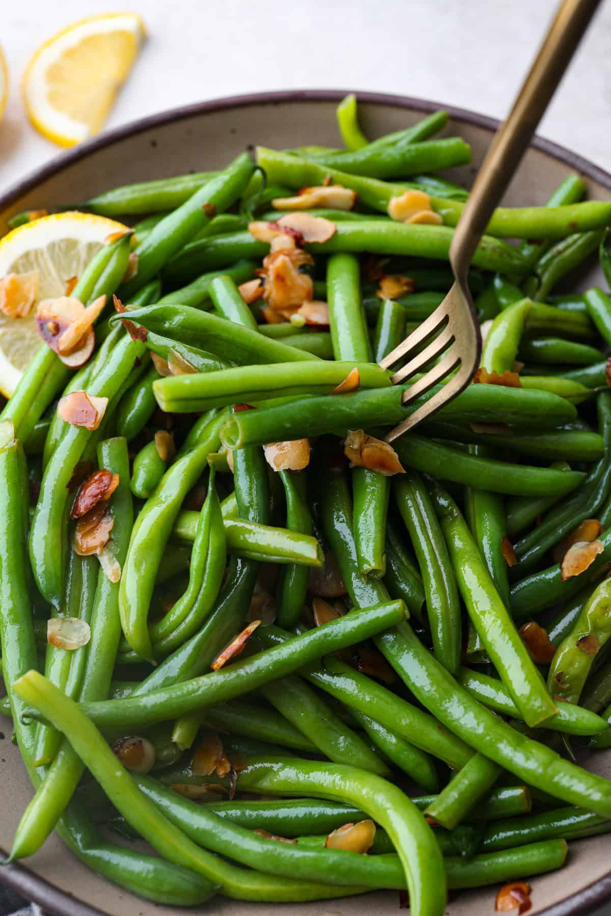 Green Beans Almondine is made with fresh green beans that are sautéed until crisp-tender and then tossed with buttery, toasted almonds. Every bite is filled with nutty richness and a hint of lemon to brighten up the flavors. Angle shot of green beans Almondine with a fork spearing some of the beans.