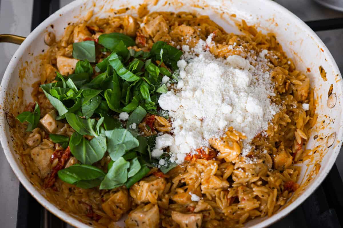 Overhead shot of basil and parmesan cheese being added to the skillet with chicken and orzo mixture. 