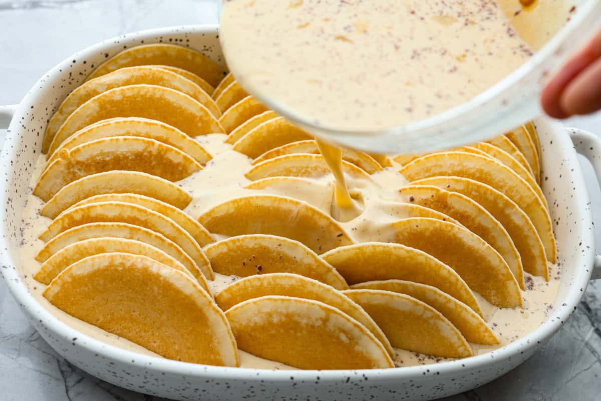 Pancake Casserole is a fun new Christmas or Easter breakfast idea. It has lots of buttery pancakes soaked overnight in a creamy custard, is baked to perfection, and served with your favorite toppings! Angle shot of custard being poured over pancakes in baking dish.