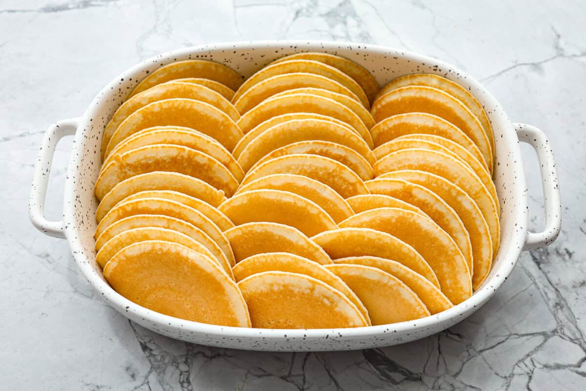 Pancake Casserole is a fun new Christmas or Easter breakfast idea. It has lots of buttery pancakes soaked overnight in a creamy custard, is baked to perfection, and served with your favorite toppings! Angle shot of pancakes stacked in baking dish.