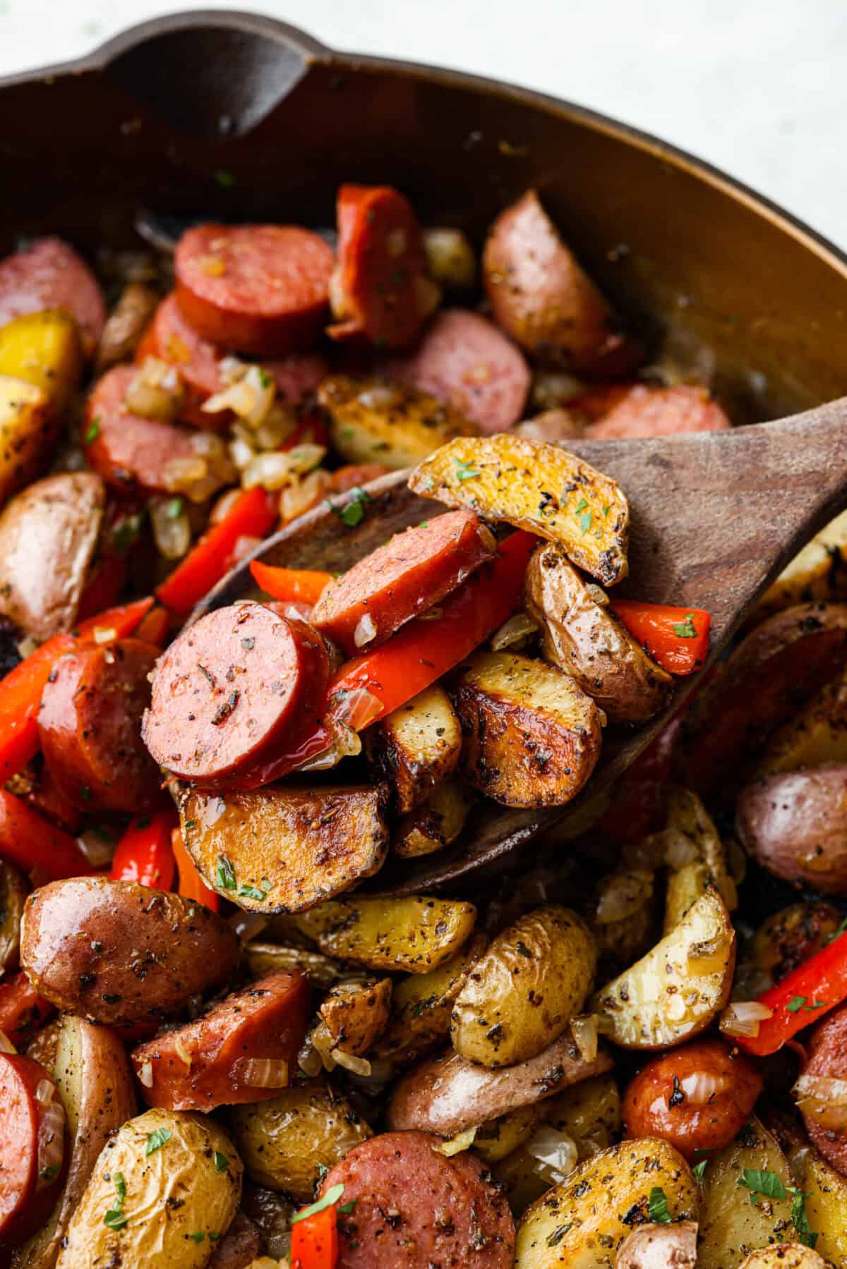 Close view of a wood spatula lifting up sausage, potatoes, and peppers.