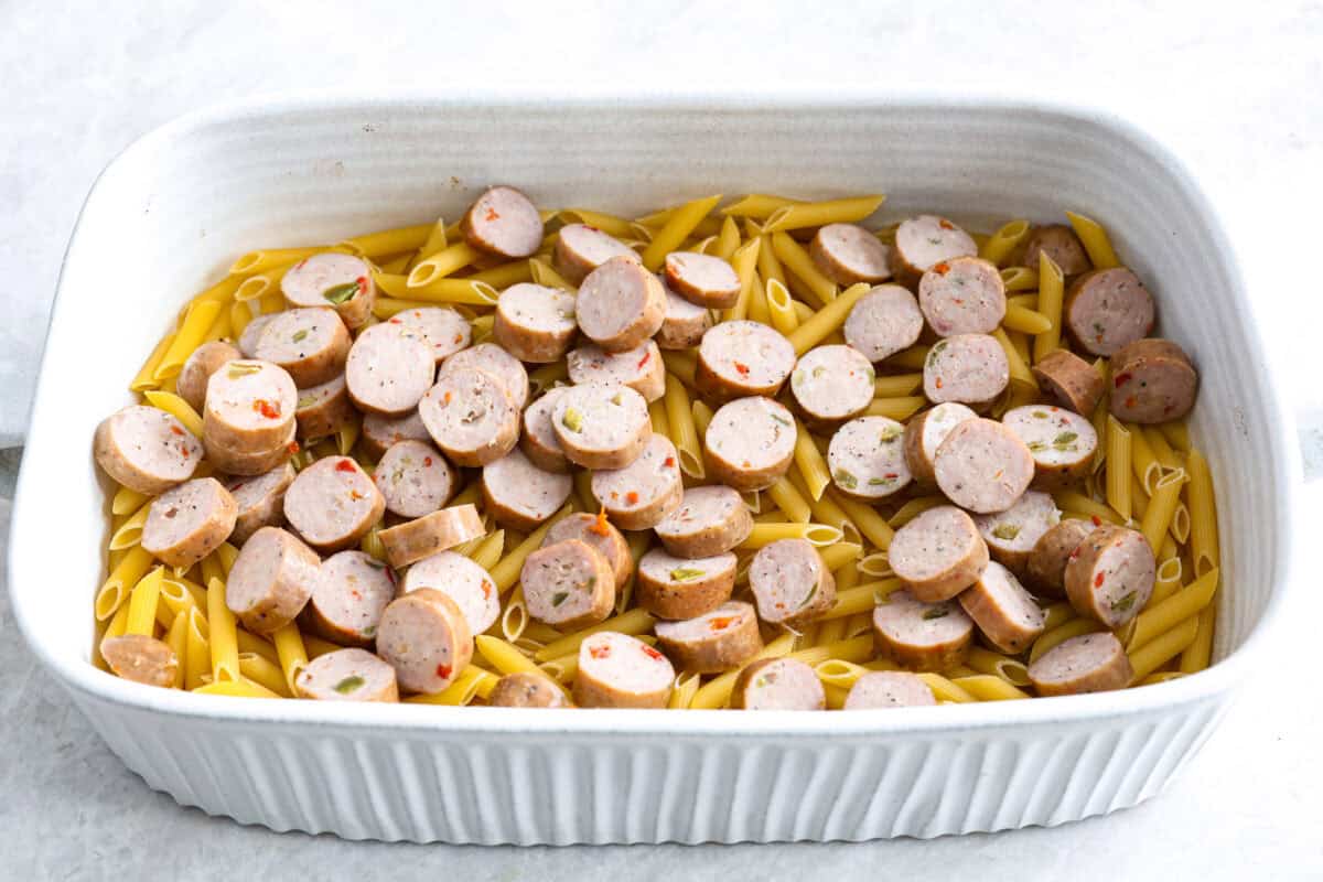 Angle shot of uncooked pasta with sausage over the top in the bottom of a baking dish. 