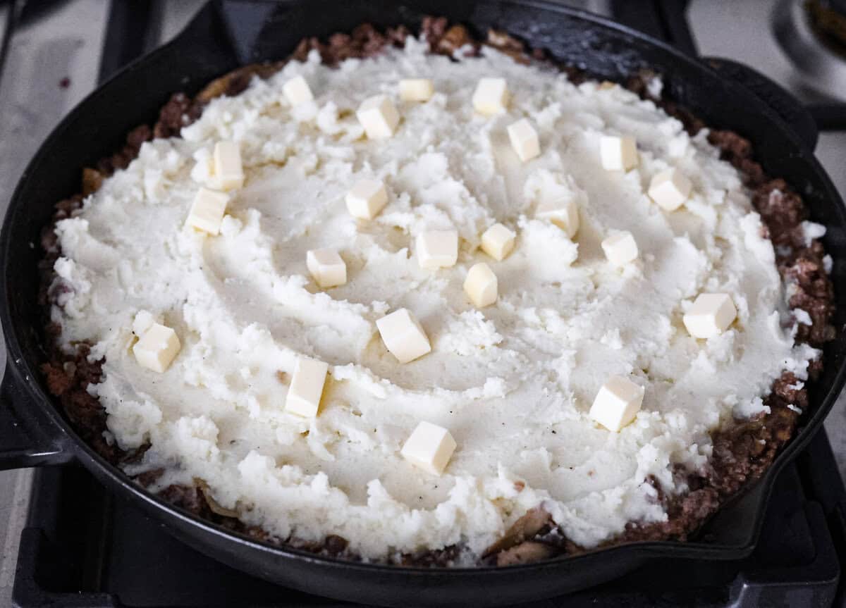 Salisbury Steak casserole is a warm and comforting savory dish. It has a thick beef gravy, tender mushrooms, and seasoned hamburger topped with rich, creamy potatoes. Angle shot of the mashed potatoes spread over the salisbury steak mixture with cubed butter on top.