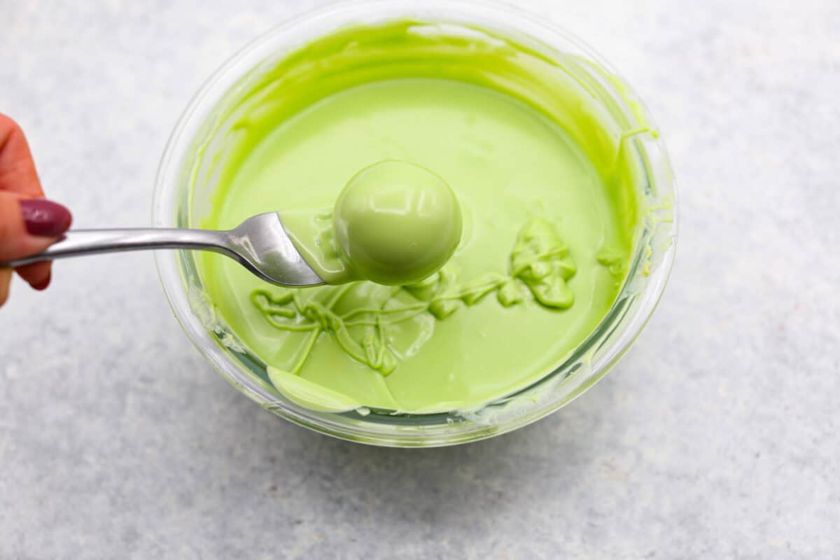 Overhead shot of green candy melt melted in a bowl with someone dipping a ball of filling in with a fork. 