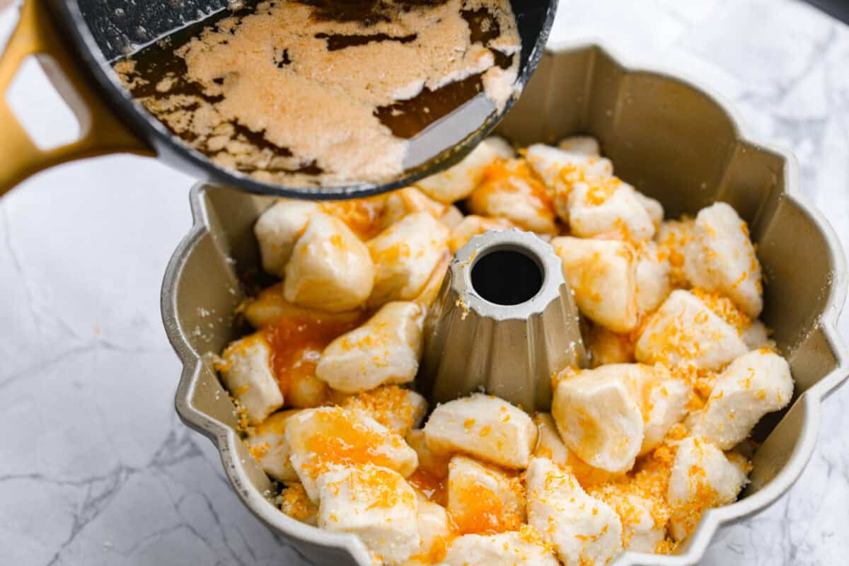 Angle shot of someone pouring the butter and brown sugar sauce over the biscuit and orange zest in the bundt pan. 