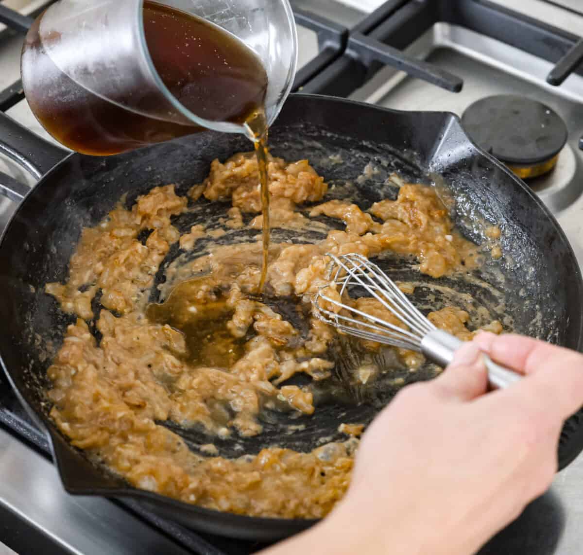 Angles hot of someone pouring beef broth into the flour and onion mixture in the skillet. 