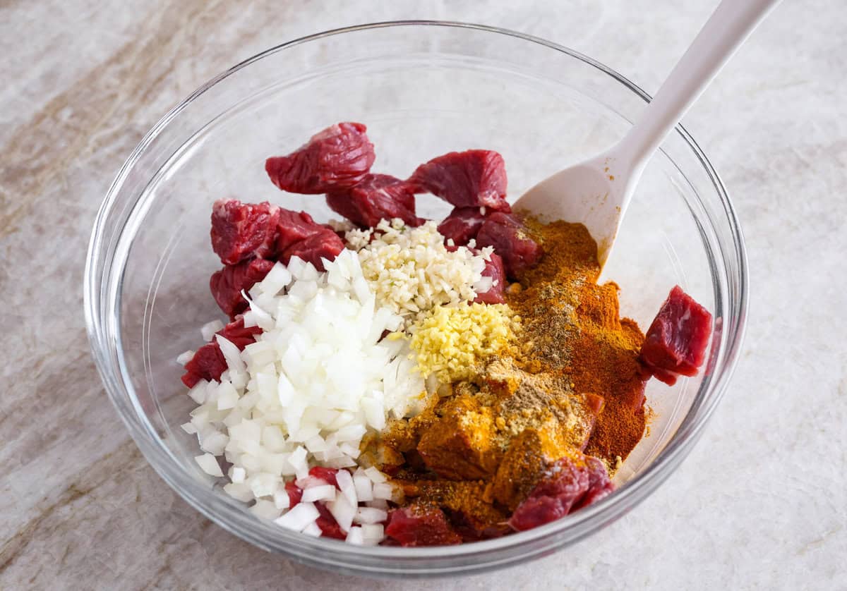 Comforting beef curry is a perfect blend of hearty and flavorful! With its mild spice level, it’s a dish that everyone can enjoy, but you can easily turn up the heat with a pinch of red pepper. Pair it with rice or naan for the ultimate cozy dinner! Overhead shot of cut up beef with diced onions seasonings, and garlic in a bowl.
