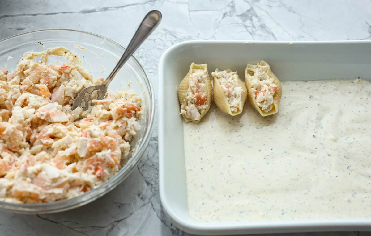 Angle shot of Alfredo sauce in the bottom of a baking dish with a few seafood stuffed shells on top of the sauce in the pan. 