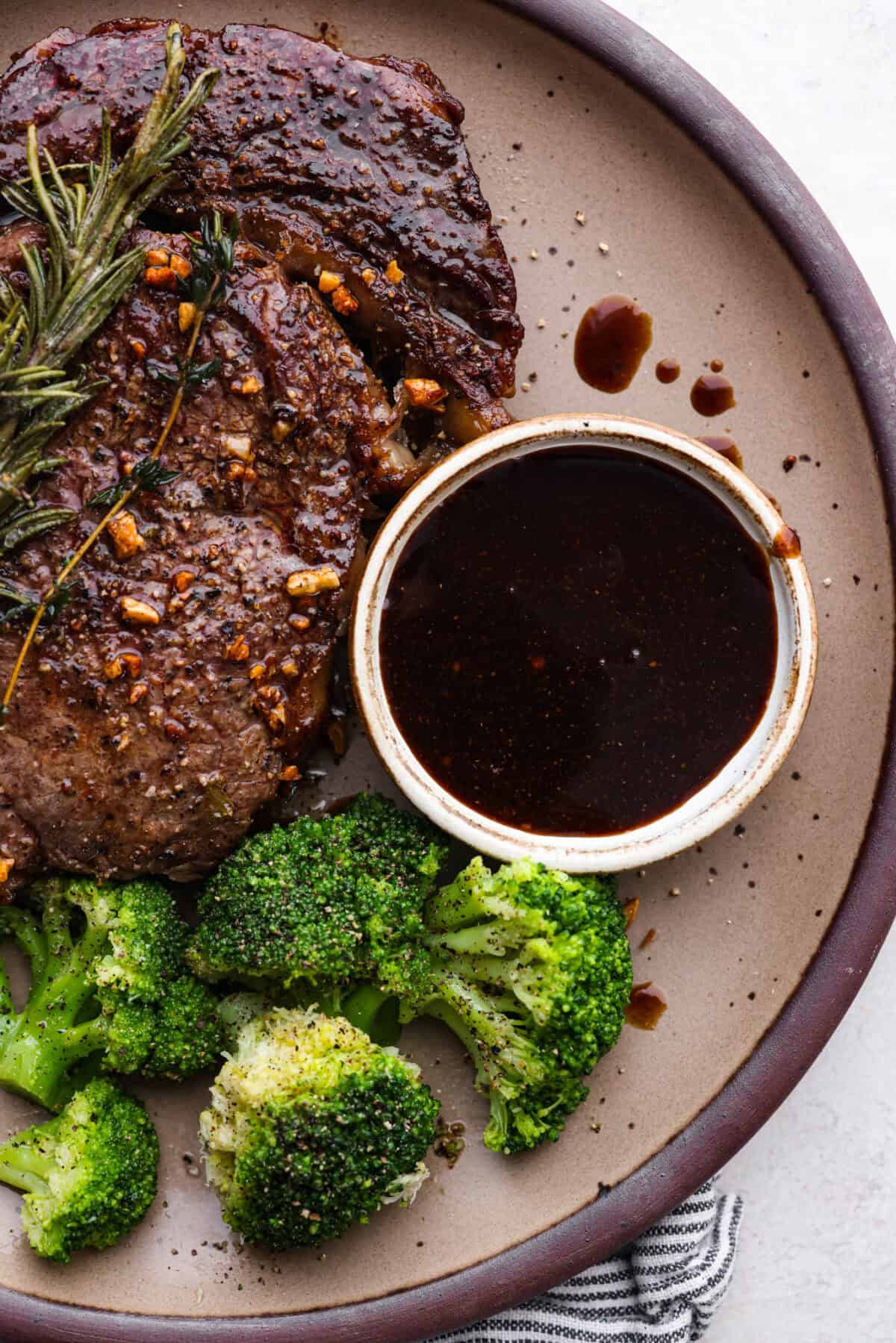 A steak shot with a steak on the table next to broccoli, and a small bowl with homemade steak sauce. 