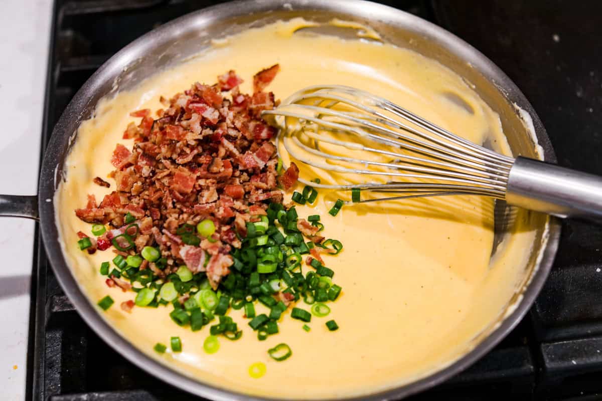 Loaded scalloped potatoes are rich, creamy, and Overhead shot of someone whisking in the bacon and green onions into the sauce.