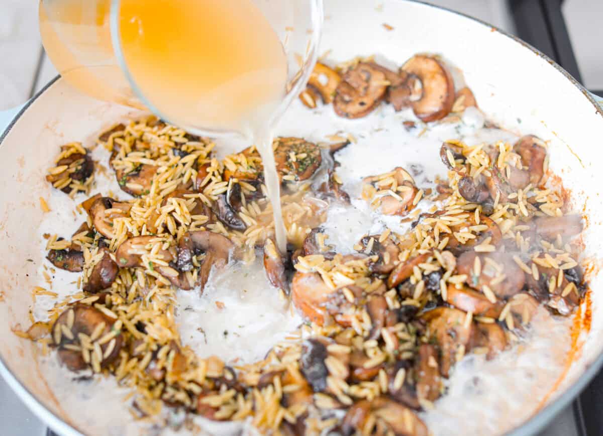 Angle shot of someone pouring cream and chicken broth in to the orzo and mushroom mixture. 