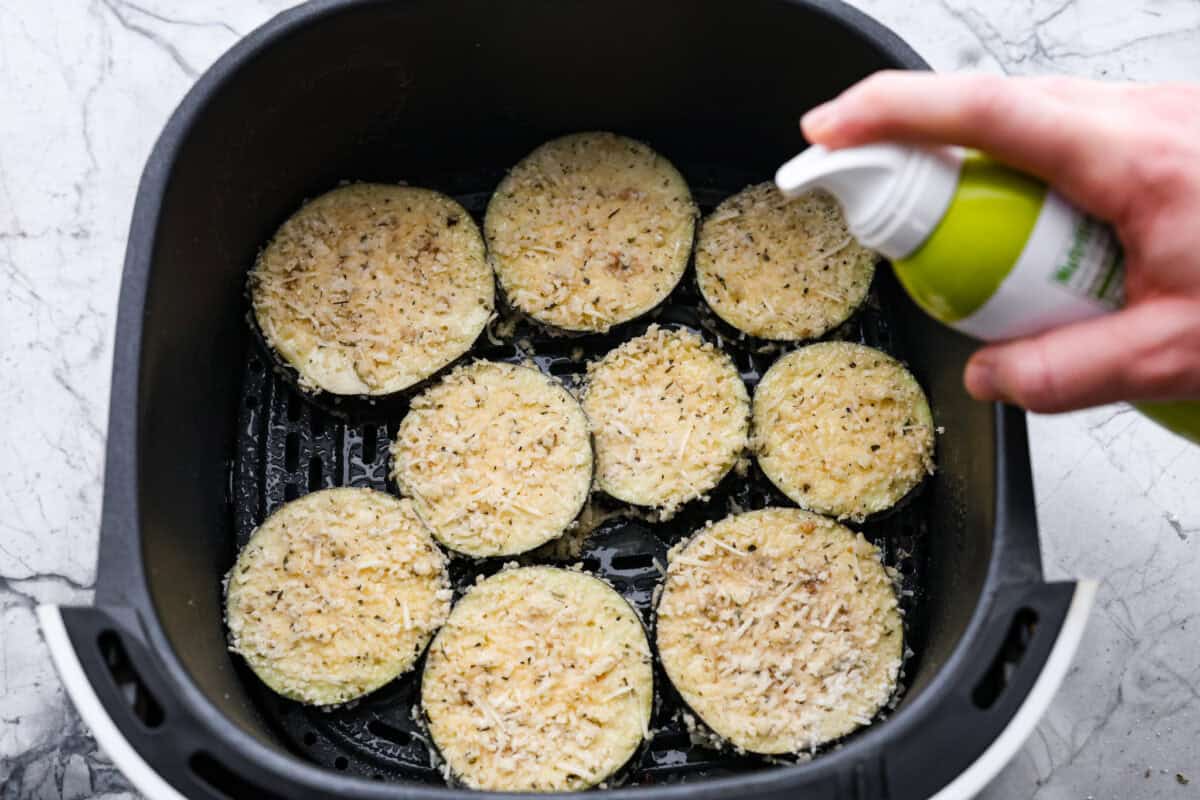 Overhead shot of someone spraying coated slices in the bottom of the air fryer basket. 