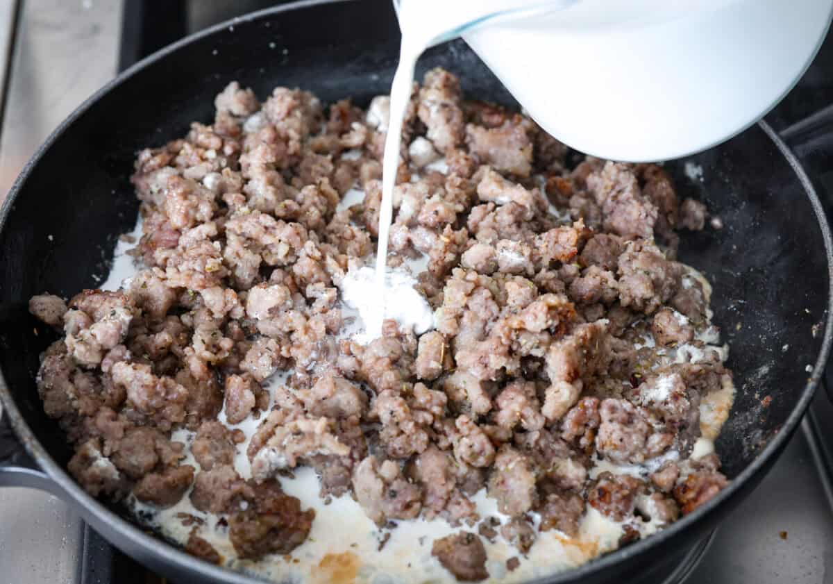 Angle shot of someone pouring half and half into a large skillet with the meat and flour mixture.