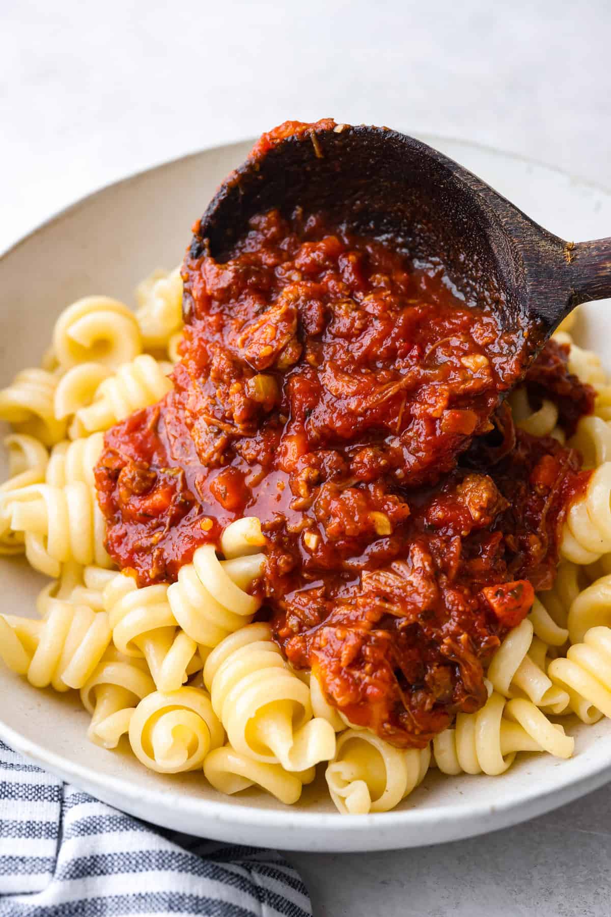 Slow cooker Sunday sauce, also known as Side shot of someone pouring a scoop of slow cooker Sunday sauce over a plate of cooked pasta.