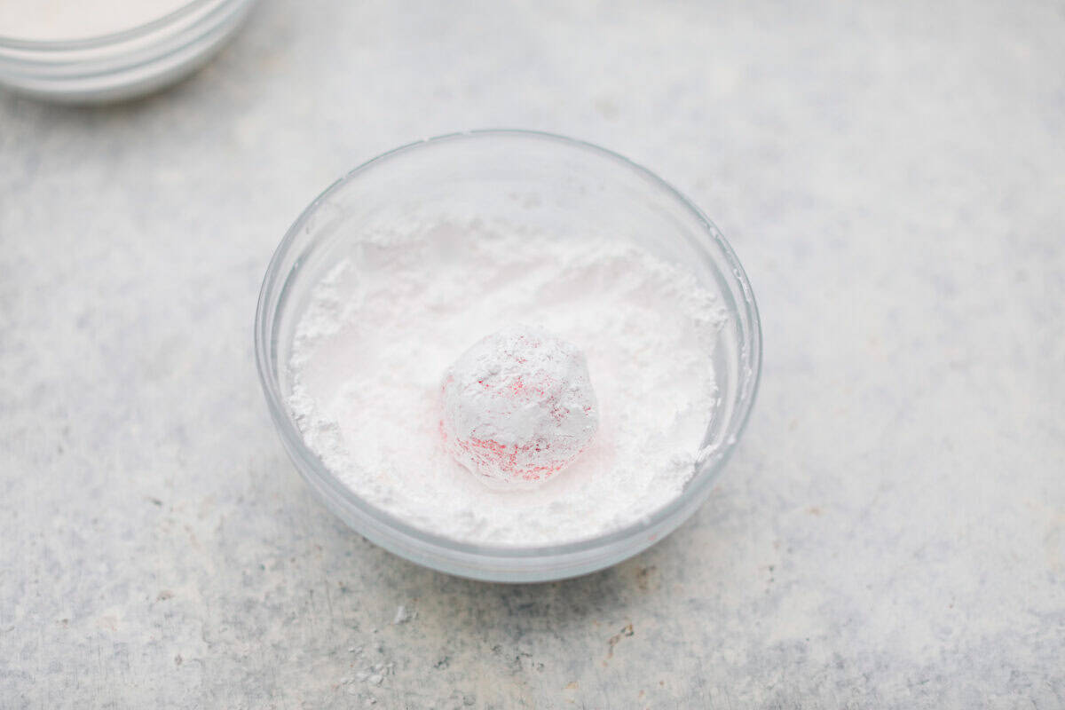 Angle shot of a ball of dough rolled in a bowl of powdered sugar. 