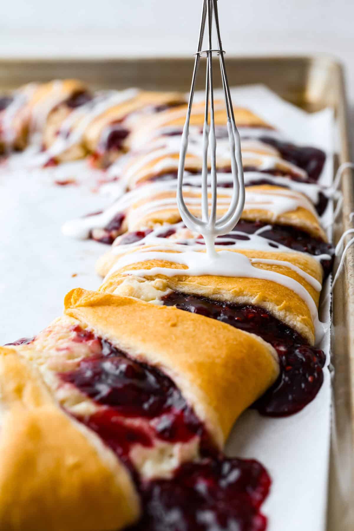 Angle shot of someone drizzling glaze over the raspberry cream cheese crescent ring with a whisk. 