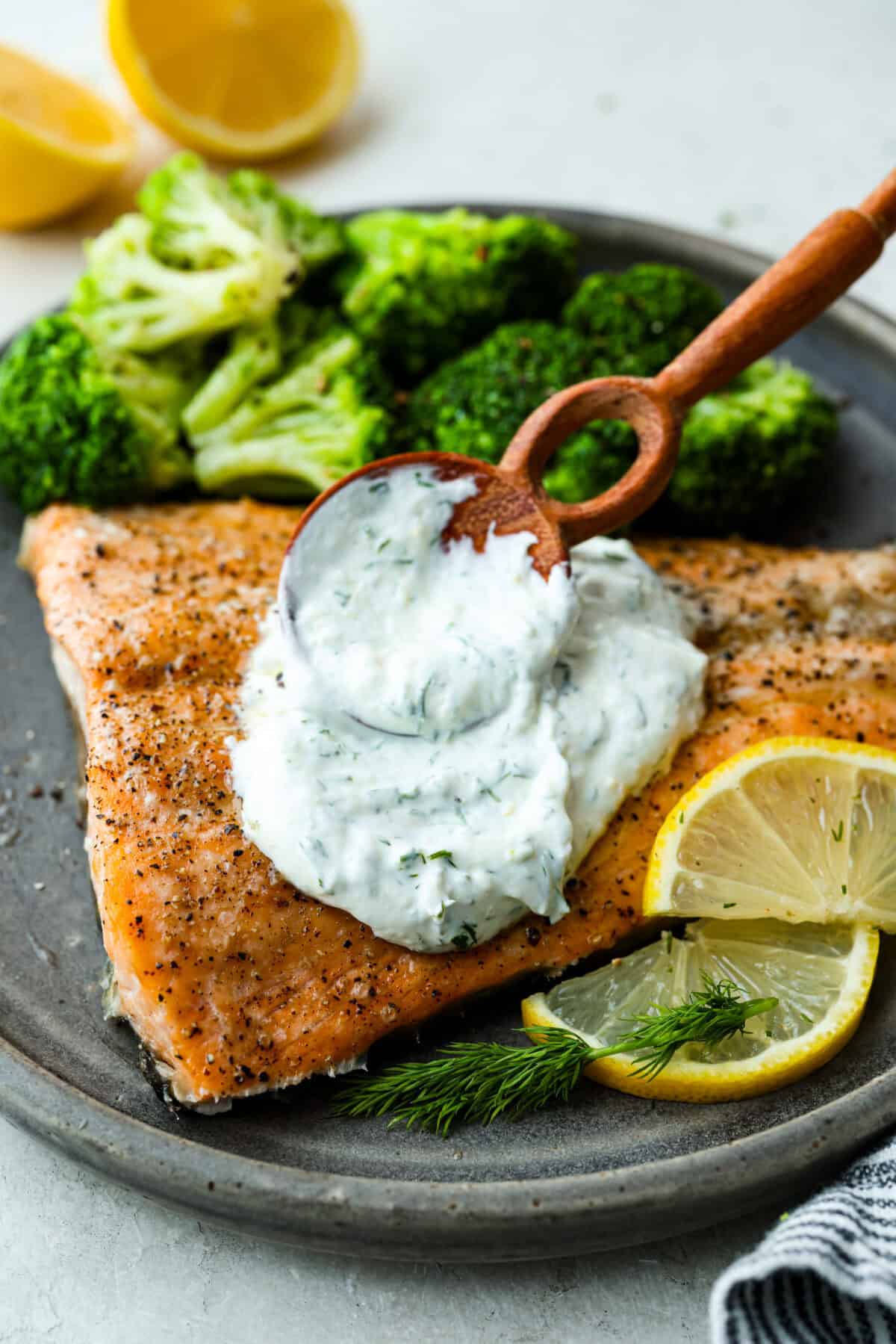 Angle shot of someone spreading fresh lemon dill sauce over a fillet of plated fish and broccoli, garnished with lemon wedges and fresh dill.