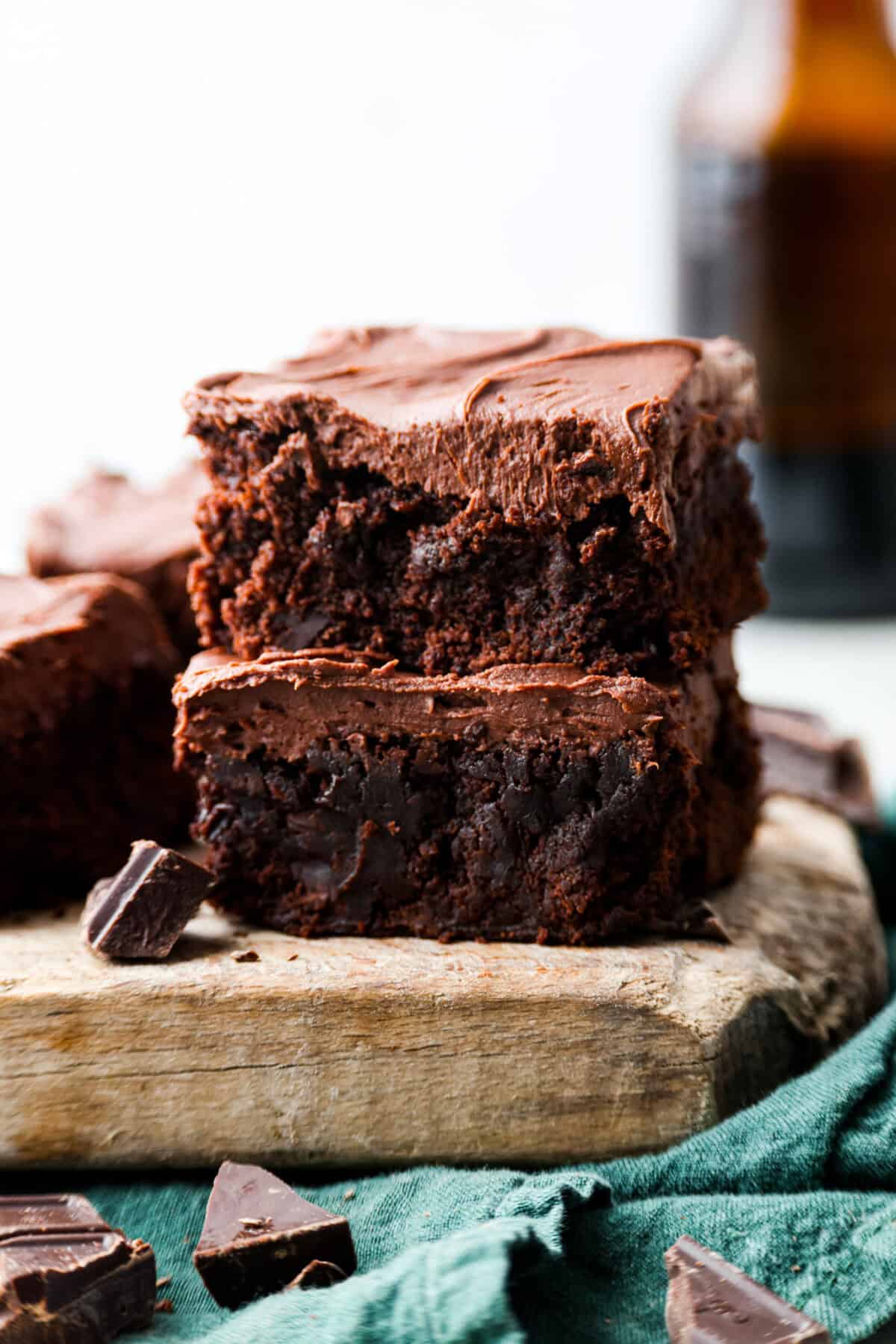 Two Guinness brownies stacked on top of each other on top of a wooden board with pieces of chocolate chunks scattered about. 