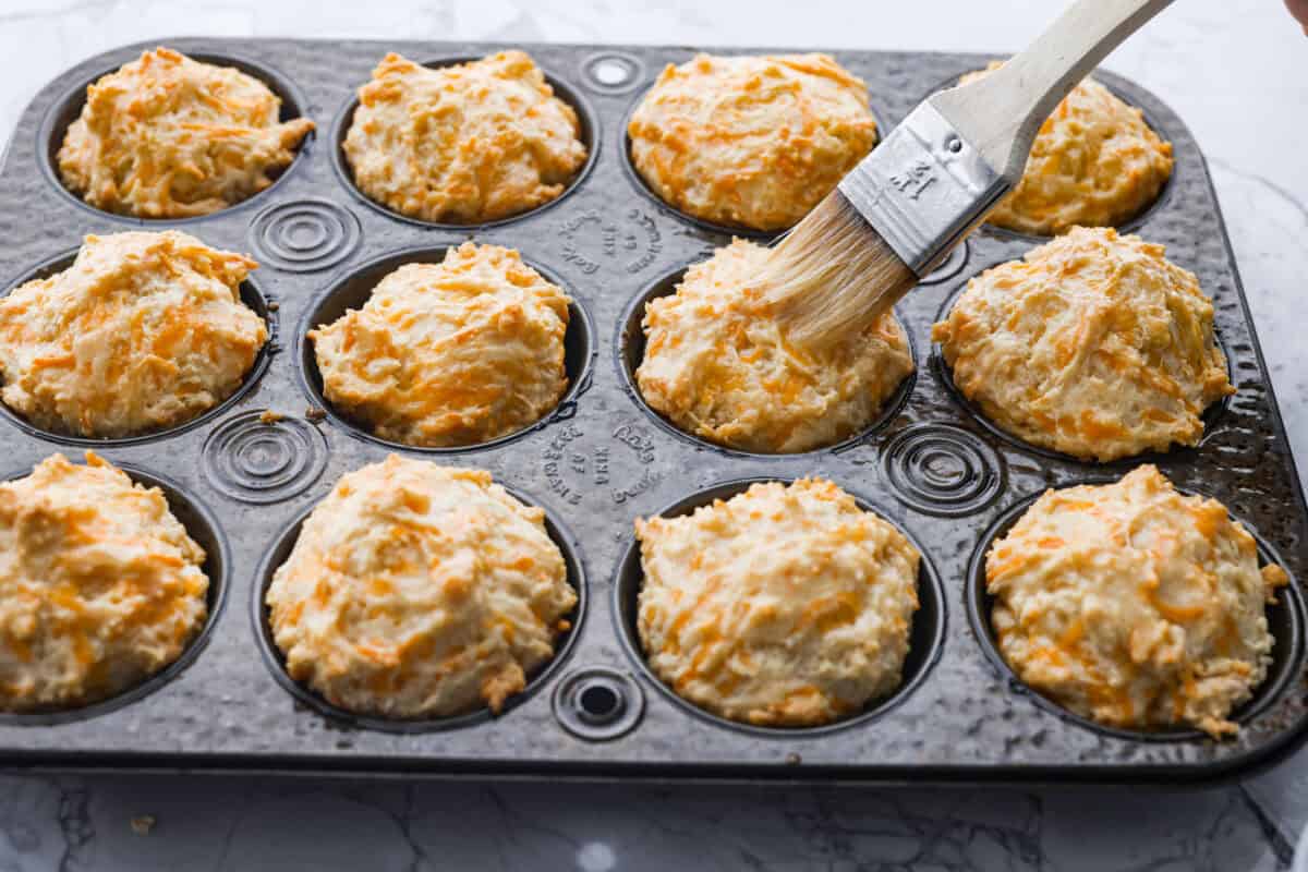 Angle shot of someone brushing butter over the top of the baked muffins. 