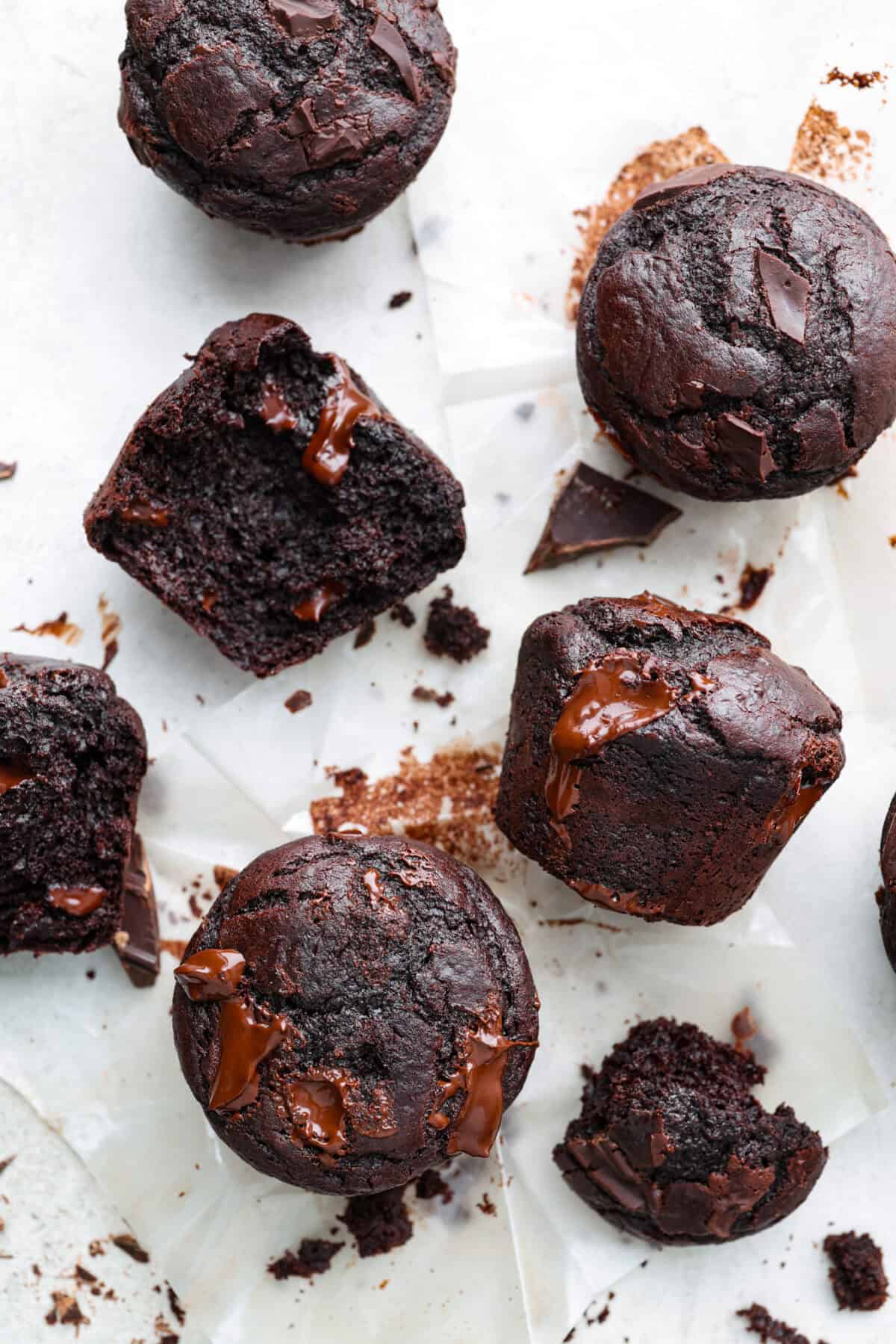 These double chocolate muffins are rich, moist, and packed with deep chocolate flavor! They are the perfect treat for breakfast, snack time, or dessert. They come together with simple ingredients and bake up beautifully with a soft, tender crumb in less than an hour! Overhead shot of double chocolate muffins scattered on parchment paper.