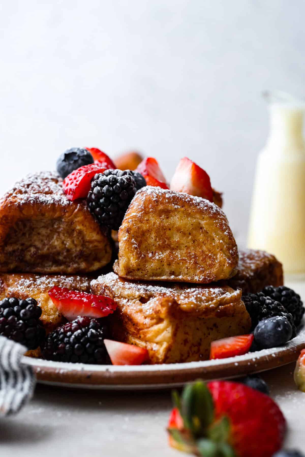 Side shot of a pile of Hawaiian roll french toast with fresh berries and a dusting of powdered sugar. 