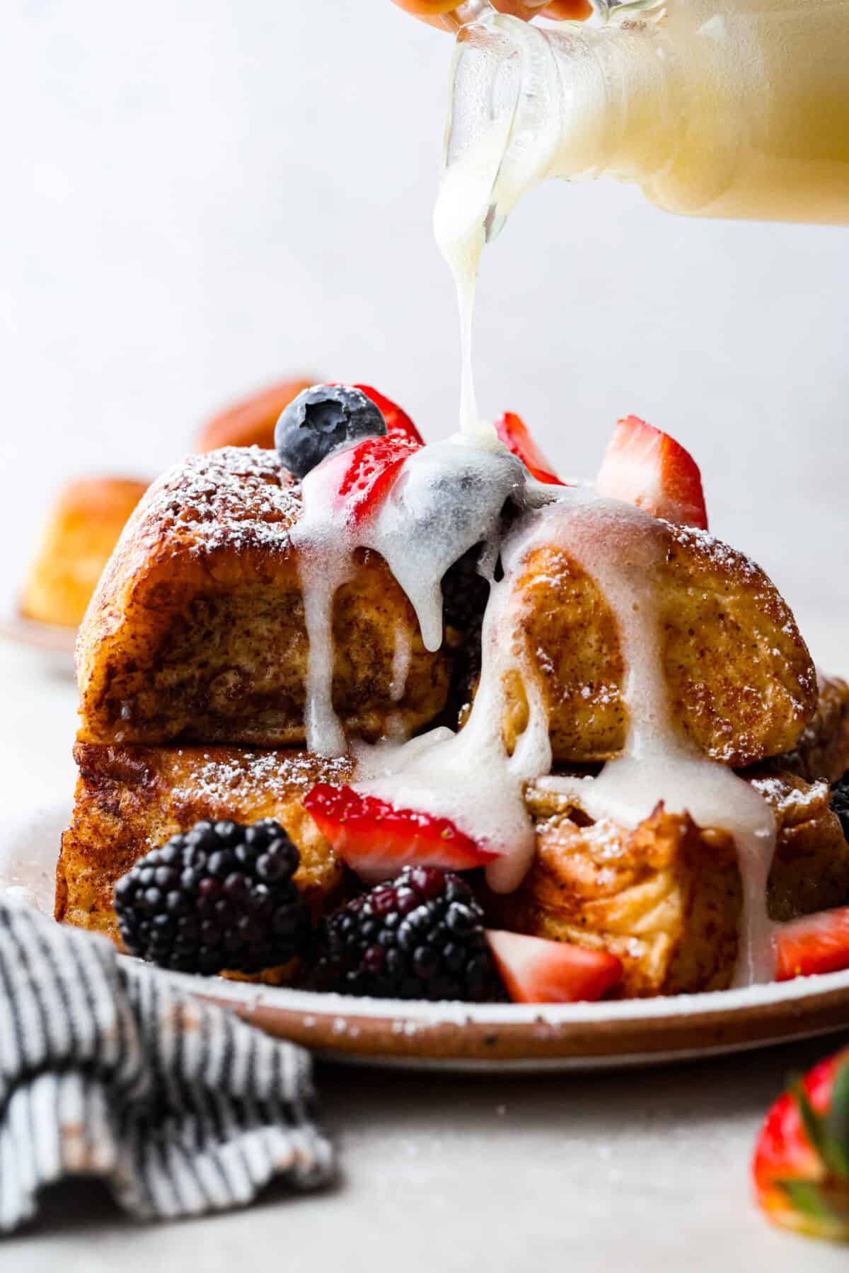 Side shot of a pile of Hawaiian roll french toast with berries and someone pouring coconut buttermilk syrup over the top. 