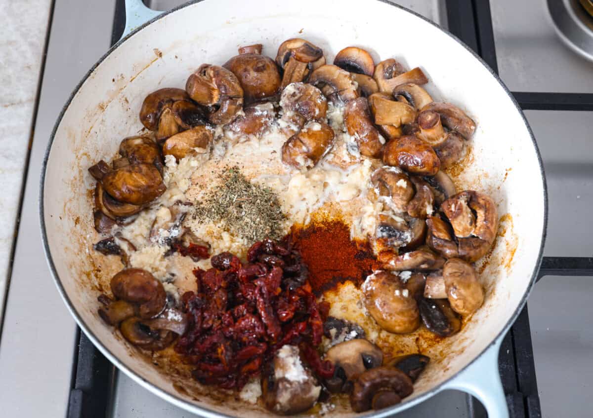 Overhead shot of the seasonings and sun-dried tomatoes adde in with the mushrooms and flour in a skillet. 