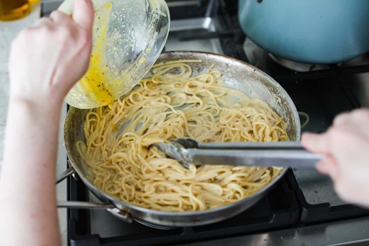 Someone pouring the carbonara sauce over the noodles in a skillet. 
