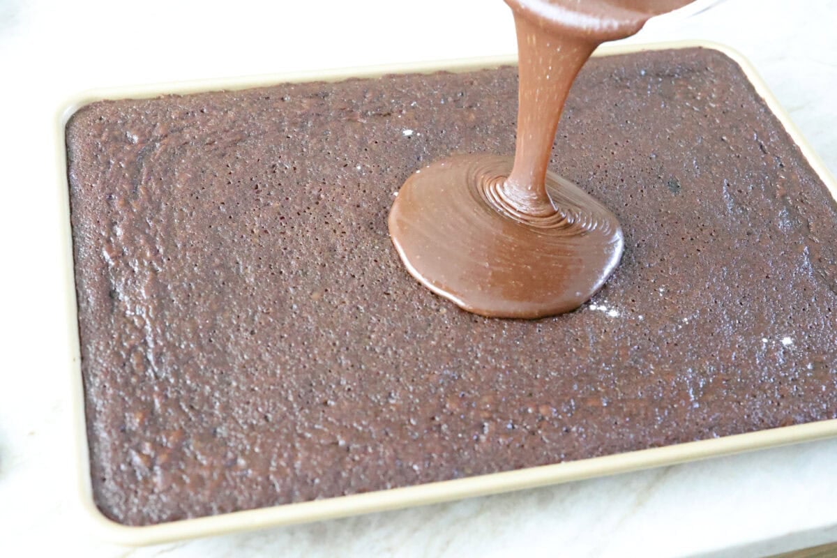 Dr. Pepper Texas Sheet Cake is what happens when your favorite fizzy drink meets rich, fudgy chocolate cake, and it’s magic! The Dr. Pepper deepens the sweetness and gives the cake this warm, almost cherry-fudge vibe that’s totally irresistible. Someone pouring the warm frosting over the warm Dr. Pepper sheet cake.