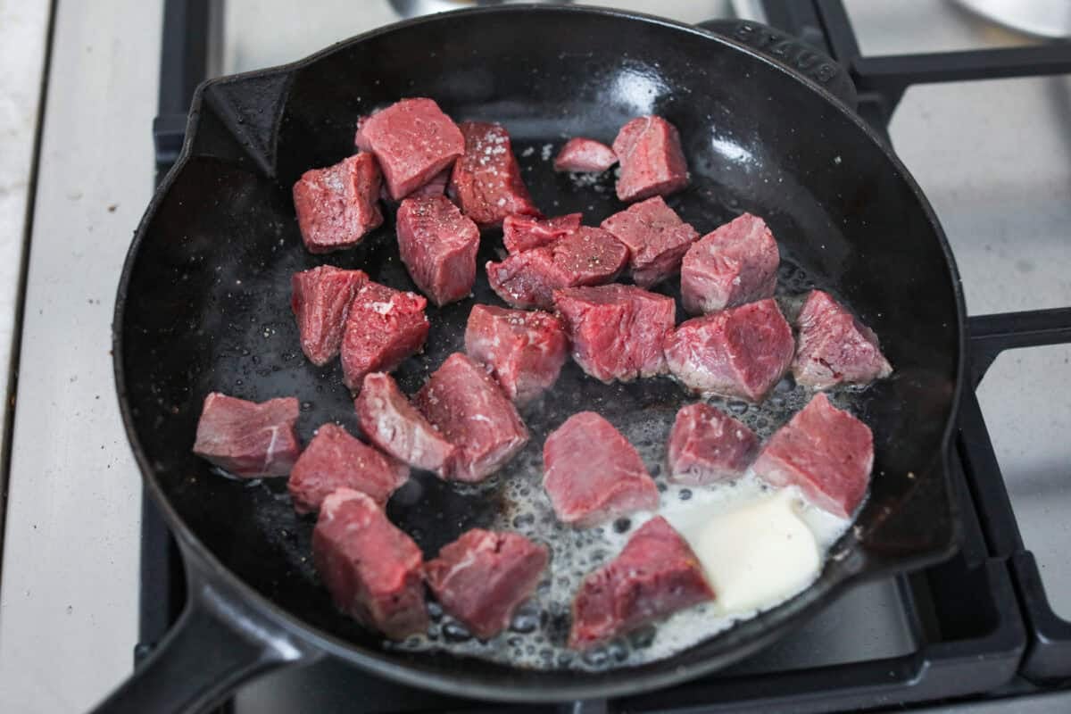 Cubed steak pieces searing in a skillet with butter melting next to them. 