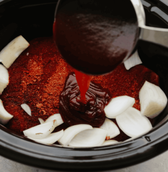 Someone pouring bbq sauce over the seasoned meat and onions in the crockpot.