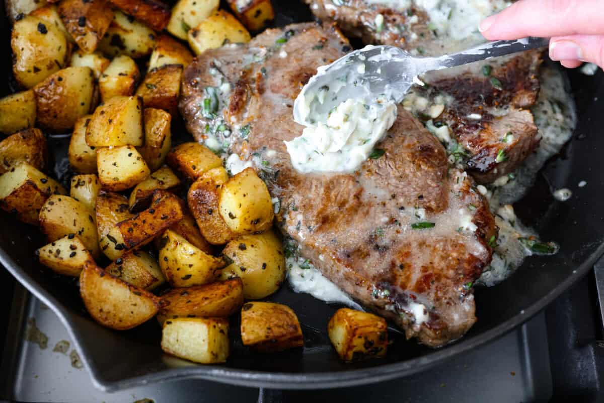 Cooked potatoes next to the cooked steaks while someone spreads the butter compound over the steak to let it melt.
