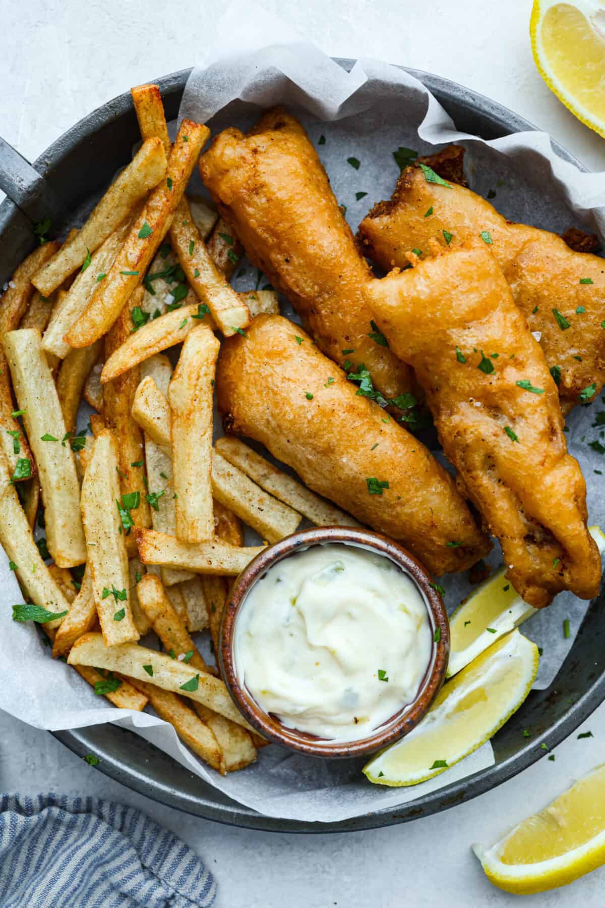 Skip the overpriced takeout and bring the pub vibes home! Crispy, golden fish and chipsโlight, flaky, pan-fried perfection. Itโs cheaper, easier, and honestly? Way better than anything stuffed in a greasy paper bag. Plate of freshly fried fish and chips with a side of tartar sauce and lemon wedges.
