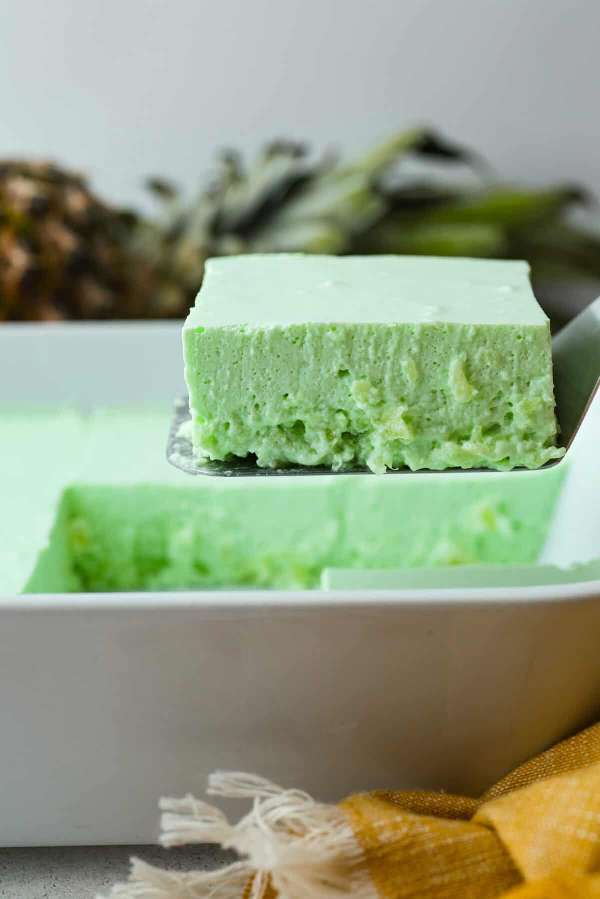 I gave the vintage lime Jello salad a modern makeover. With cream cheese for tang, pineapple and lime for zip, and a splash of 7-Up for that light, fizzy fluff, this retro-inspired treat is now a sweet, citrusy dream I know you guys will love! Someone lifting a slice of lime jello salad out of the dish.