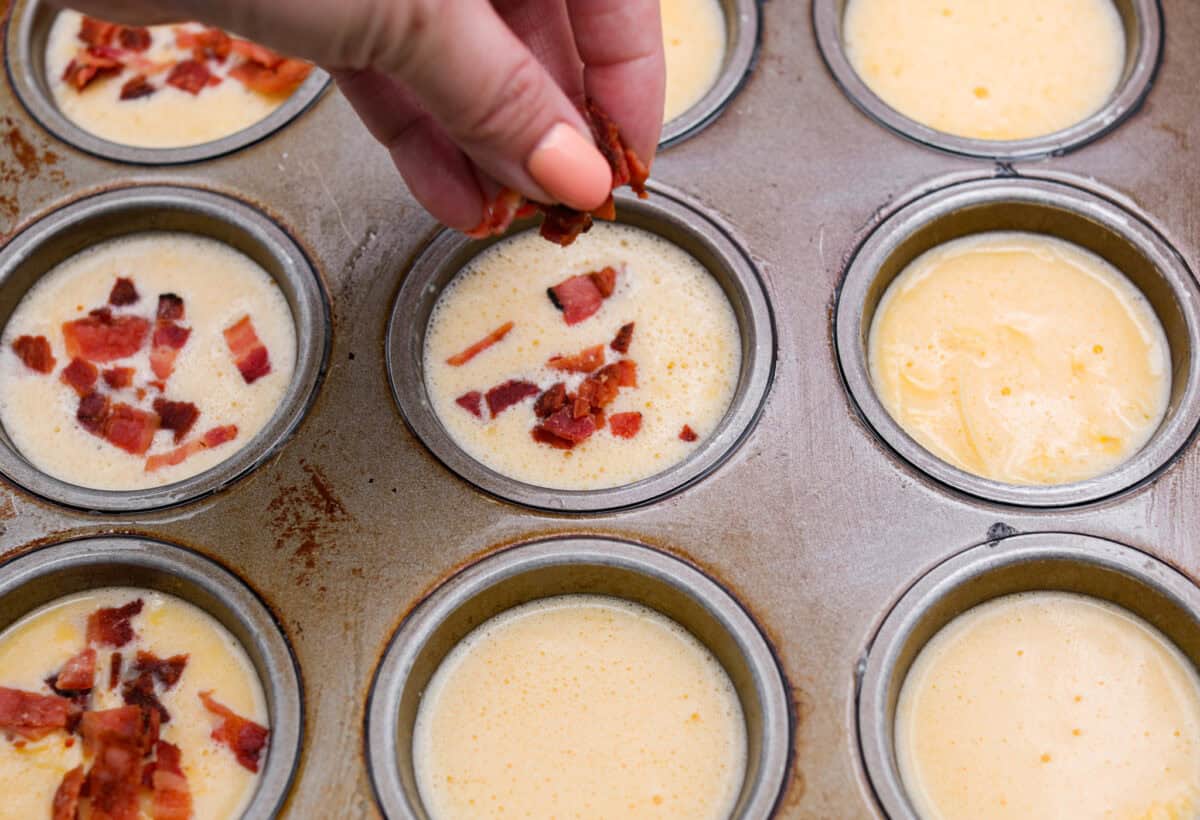 I’ve been hooked on Starbucks egg bites for years, but not the price! This homemade version is just as fluffy and flavorful, and makes the perfect grab-and-go, protein-packed breakfast. Someone sprinkling bacon over the divided egg mixture in the muffin tins.