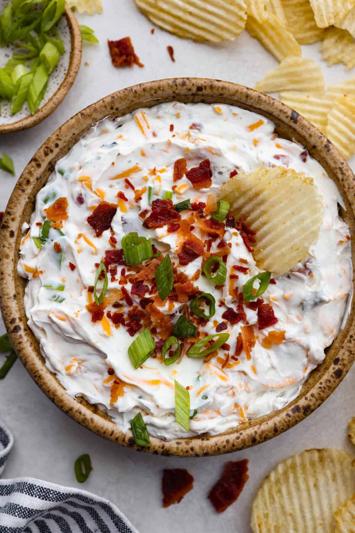 Creamy, cheesy, and loaded with crack dip flavor! This 6-ingredient appetizer is served cold and ready in just 10 minutes. Good luck keeping it on the table. Overhead shot of a serving bowl with crack dip in it garnished with cheese, bacon, and green onions, with a ruffled chip sticking out.