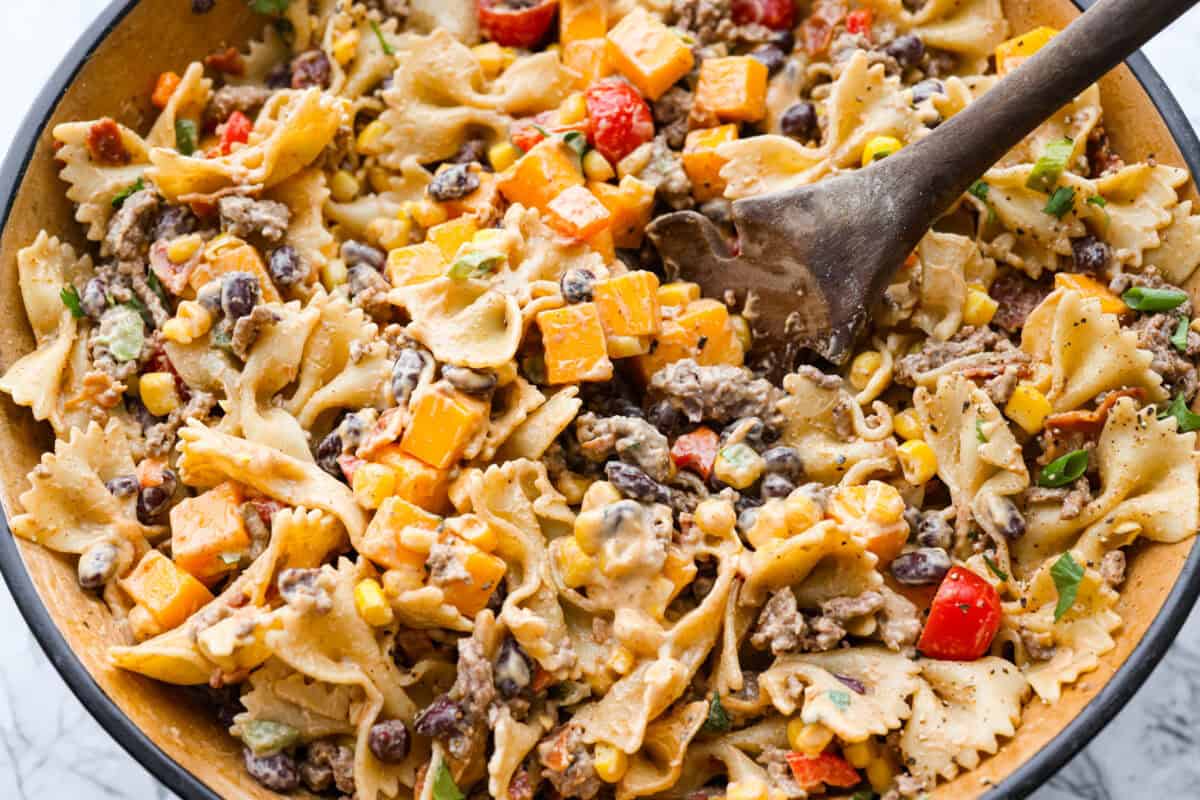 Yeehaw! My cowboy pasta salad is loaded with beef, bacon, beans, and all the fixings for all you cowboy lovers. You'll get all the flavors of the Wild West tossed in the tastiest cowboy salad dressing. Someone stirring together the pasta salad ingredients with the dressing.