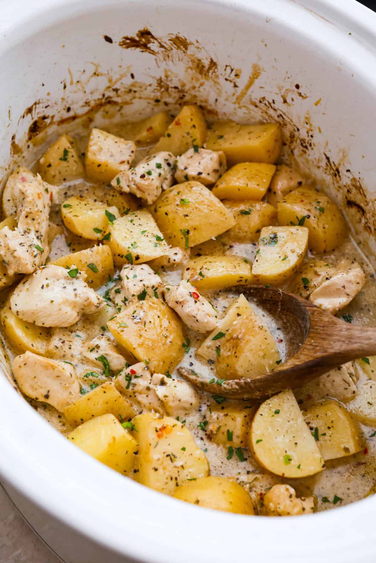 This viral slow cooker Parmesan garlic chicken has juicy meat, tender potatoes, and a sauce so rich and creamy that scooping it with a spoon won’t cut it. You’ll want to drink it, or let’s be real, bathe in it. Overhead shot of cooked slow cooker parmesan garlic chicken and potatoes with wood spoon.