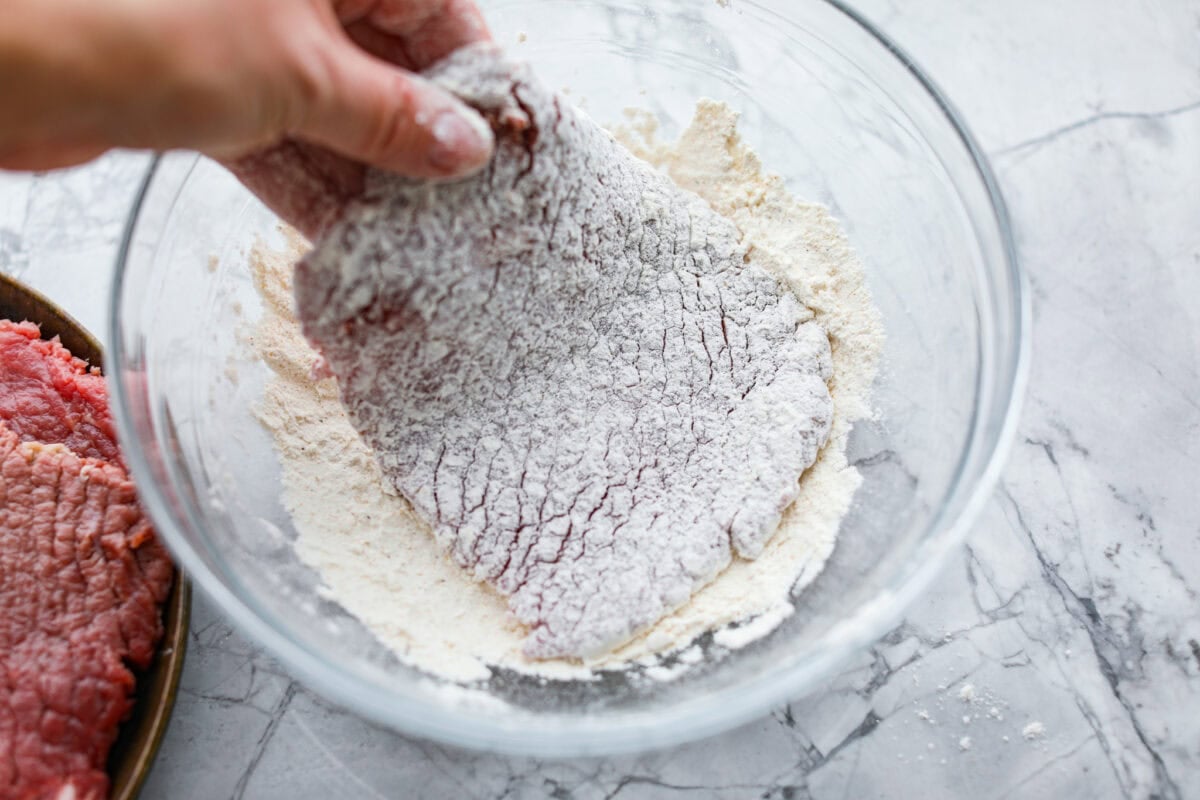 Cube steaks are my go-to budget comfort food. They're quick, cozy, and smothered in gravy. I love them with mashed potatoes or veggies, and I’ve got a simple trick to keep them melt-in-your-mouth tender every time. Someone dipping the smashed steak into the breading mixture.