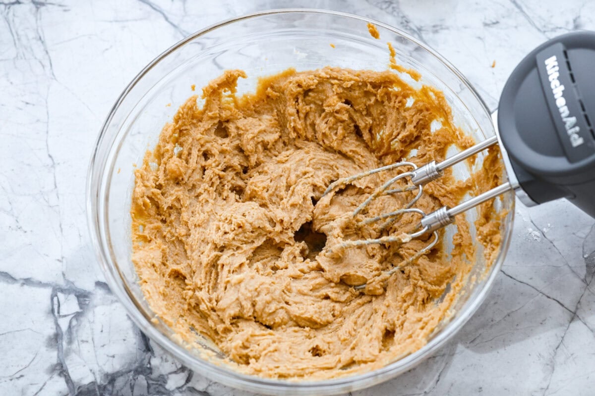 Someone mixing butter, brown sugar, and peanut butter in a bowl with a hand mixer. 